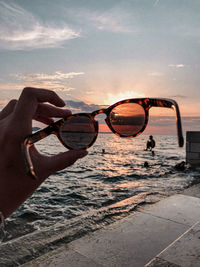 Hand holding sunglasses at beach against sky during sunset