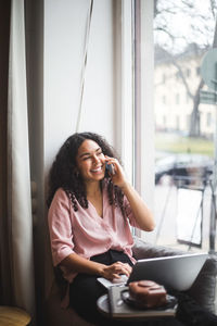 Smiling young female entrepreneur talking through smart phone sitting on window sill in office