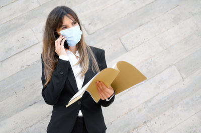Businesswoman wearing mask talking on phone standing at against wall