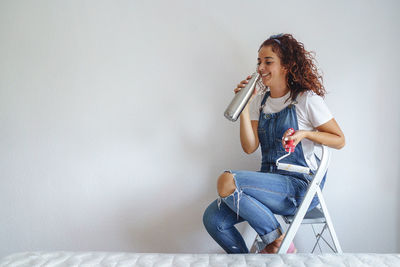 Smiling woman drinking water while sitting against wall