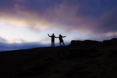 Silhouette of man and woman with arms outstretched while standing on landscape against sky during sunset
