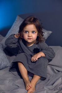Girl in a gray dress sits on a bed with pillows in a bedroom with a blue wall