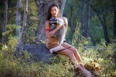 Smiling young woman in traditional clothing sitting with raccoon in forest