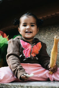 Portrait of happy girl holding rolling pin while sitting against built structure