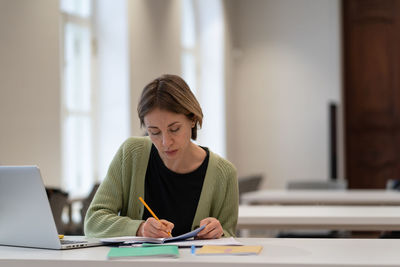Middle-aged female student preparing for exam in public library while sitting at table with laptop