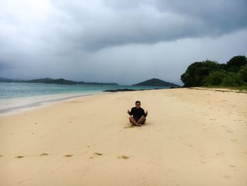 Man sitting on sand against cloudy sky at beach