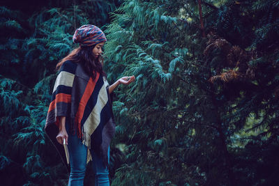 Side view of woman with book touching tree branch while standing in forest