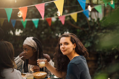 Portrait of confident young woman holding drink while sitting at table during garden party