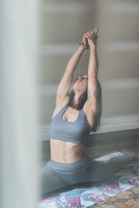 Through glass view of flexible female in sportswear stretching legs and looking up with raised arms while practicing yoga in house