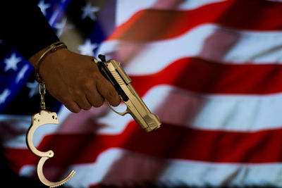 Cropped hand of male criminal holding gun against american flag