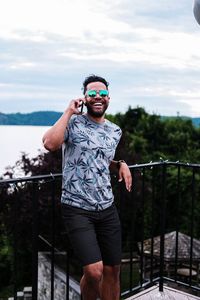 Young man wearing sunglasses standing by railing against sky