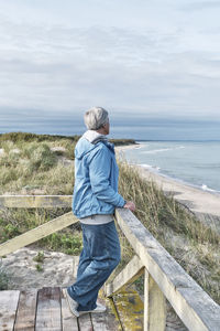 Senior woman with short gray hair in blue jeans and windbreaker standing on wooden platform by sea.