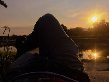 Rear view of man sitting by river against sunset sky