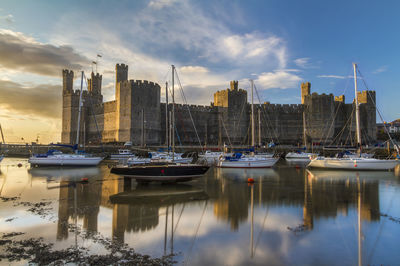 Boats moored in harbor by castle during sunset