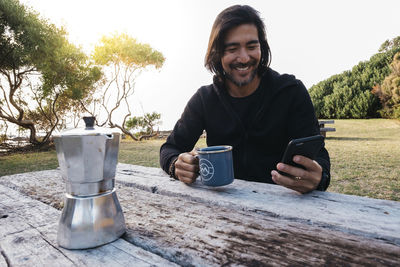 Happy man with coffee using mobile phone on wooden table while sitting against sky