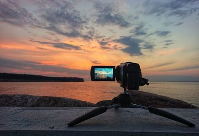 Camera on tripod by sea against sky during sunset