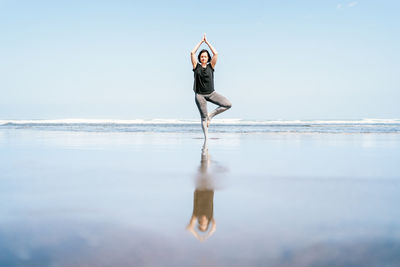 Full body young female in sportswear standing in tree pose with arms up while practicing yoga on sandy beach against waving sea with eyes closed
