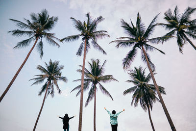 Rear view of friends with arms outstretched standing amidst palm trees against sky