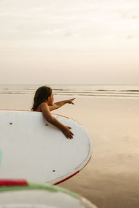 Girl with paddleboard pointing at sea on beach during sunset