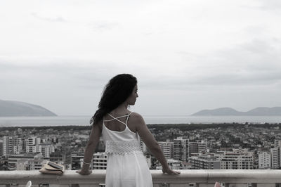 Rear view of woman standing by railing while looking at cityscape against sky