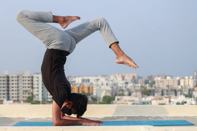 Side view of man doing yoga on highrise building terrace against sky