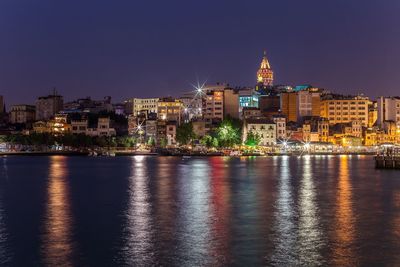 Illuminated galata tower amidst buildings in front of river against clear sky at night