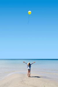 Rear view of girl standing with arms outstretched standing on shore at beach against blue sky