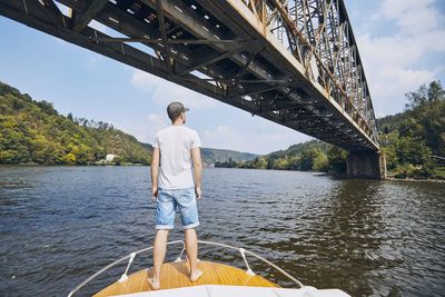 Rear view of man standing on boat in river below bridge