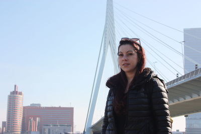 Portrait of young woman standing against bridge and sky in city