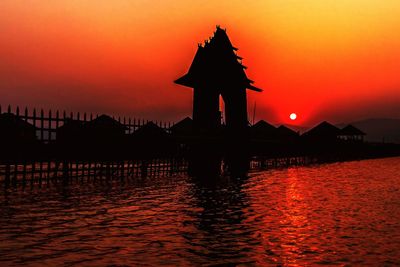 Silhouette building by sea against orange sky