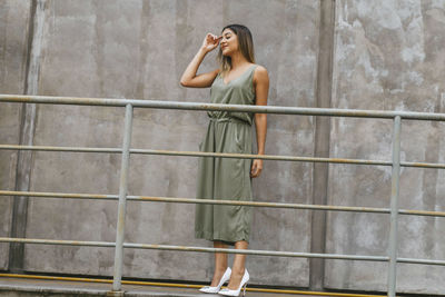 Full length of fashionable young woman standing by railing against concrete wall