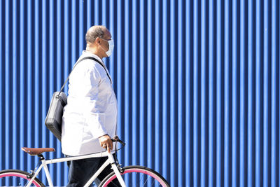 Mature man walking with bicycle by blue metal wall on sunny day