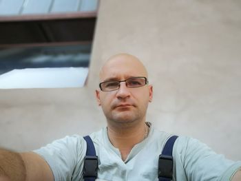 Low angle portrait of bald man wearing eyeglasses while sitting against wall