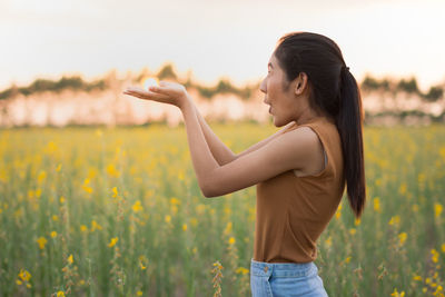 Optical illusion of young woman holding sun while standing on oilseed rape field against sky during sunset