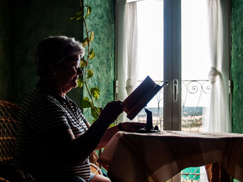 Woman reading book while sitting by window at home