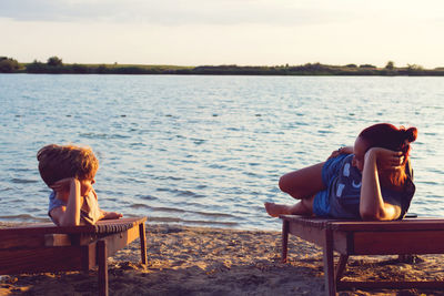 Mother and son lying on lounge chairs at beach against sky