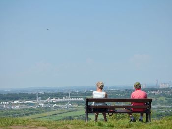 Rear view of man and woman sitting on park bench against clear sky during sunny day