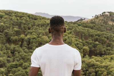 Back view of pensive african american male in trendy clothes standing in nature and admiring view of mountainous forest