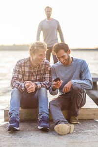 Happy male friends using mobile phone on pier with man walking in background