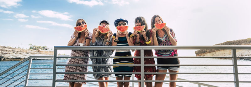 Portrait of friends eating watermelon while standing against sky