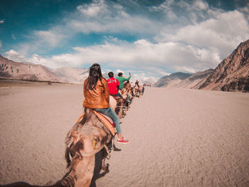 Rear view of people riding camels on desert against sky
