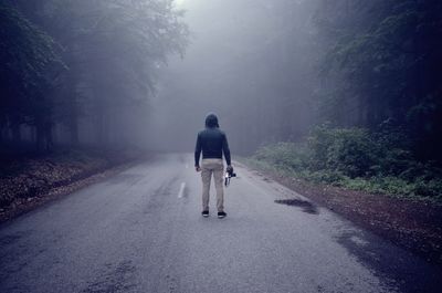 Rear view of man holding camera while standing on road amidst trees during foggy weather