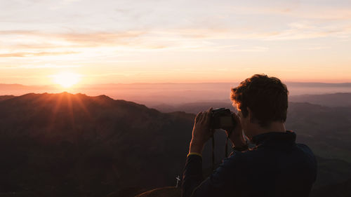 Rear view of young man photographing on mountain against sky during sunset