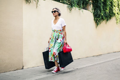 Fashionable woman walking on footpath with bags against wall
