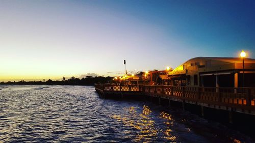 Illuminated buildings by sea against clear sky at sunset