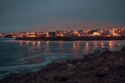 Illuminated buildings by sea against sky at dusk