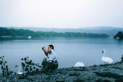 Side view of woman photographing while crouching at lakeshore