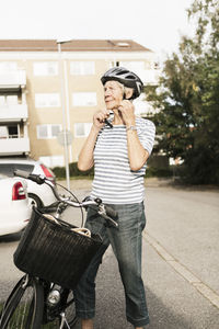 Smiling senior woman wearing helmet while standing by bicycle against house