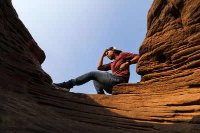 Low angle view of young man sitting on rock formation against sky