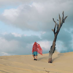 Woman standing by dead tree at desert against cloudy sky